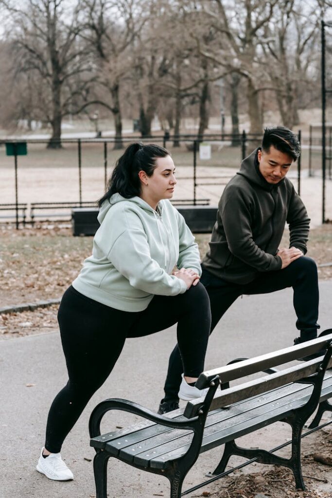 Side view of plus size woman with Asian male personal trainer in sportswear stretching legs on bench during outdoor workout in city park